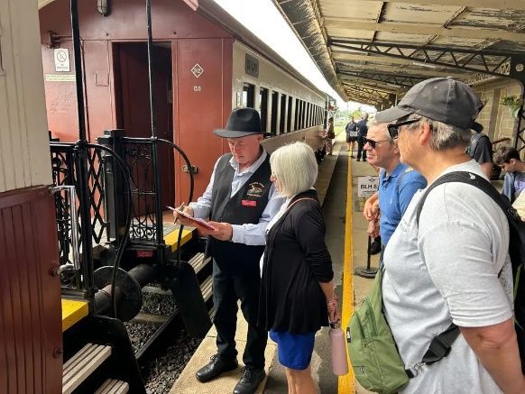 People waiting beside a train platform as a conductor checks tickets beside a maroon carriage