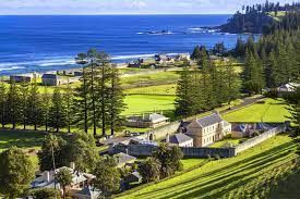 Coastal hillside with green fields, houses, tall trees, and blue ocean in the distance