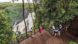 Wooden viewing deck beside a tall waterfall in a lush green forest, with a few visitors standing and sitting nearby