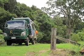 Green truck driving on a grassy dirt path beside a forest, with a wooden fence post in the foreground