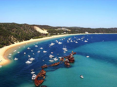Aerial view of a curved sandy bay with many sailboats and a rusted shipwreck near the shore