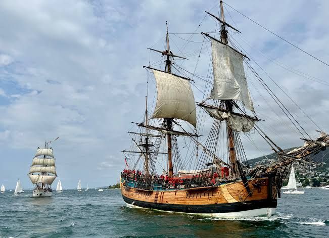 Tall sailing ship with white sails on calm water, with other ships in the background under a cloudy sky