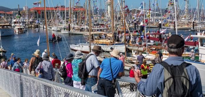 Crowd walking along a harbor dock beside moored fishing boats and sailboat masts under a clear blue sky