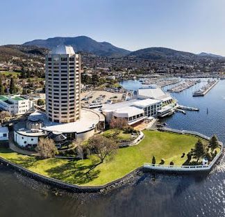 Harborfront marina with a tall hotel tower, docks, and green waterfront lawn beneath mountains