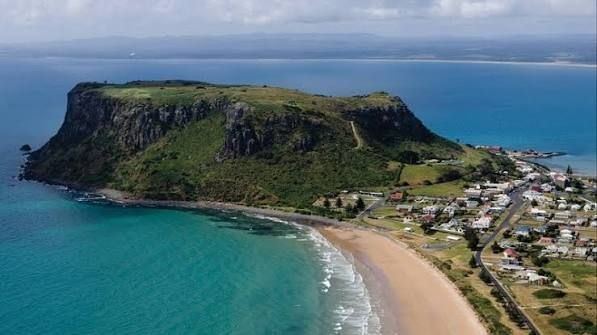 Aerial view of a green coastal headland beside a turquoise bay and sandy beach with a seaside town