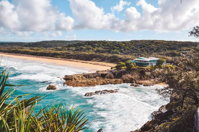 Rocky turquoise cove with sandy beach, ocean waves, and a small house on a coastal cliff under cloudy sky