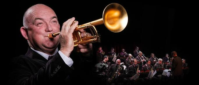 A man in a suit playing a trumpet, with a small orchestra lit in the background.