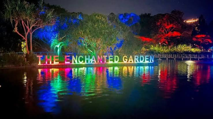 Colorful illuminated sign reading “THE ENCHANTED GARDEN” reflected on a calm lake at night