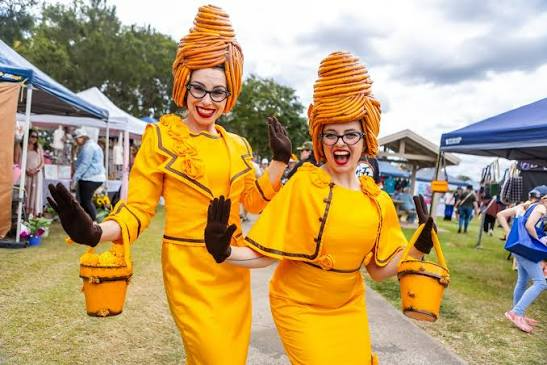 Two people in bright yellow costumes and tall orange hats smiling at an outdoor fair
