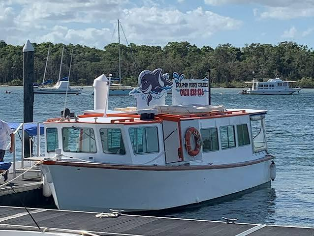 White tour boat with orange trim docked on a calm harbor, with a sign on top and forested shoreline in back