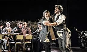 Actors performing onstage, one man embracing another, with seated audience members visible behind them.