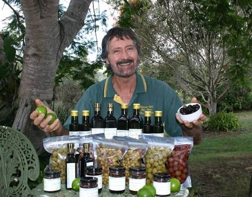 Man outdoors by a table displaying bottles and jars of preserved food, holding a lime and a bowl of berries