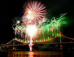 Fireworks bursting over a lit bridge at night, with red, green, and white reflections on the water
