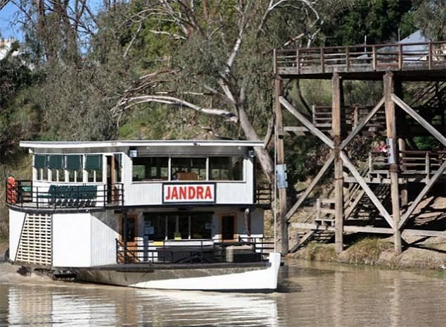 White riverboat named JANDRA beside a wooden riverside structure on muddy water