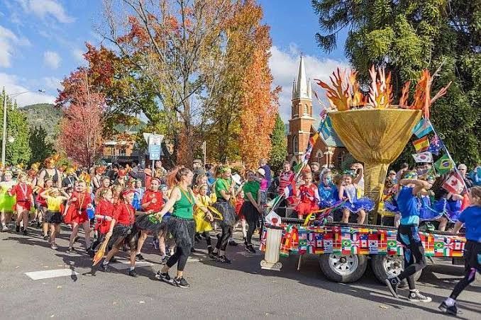 Colorful parade dancers in bright outfits moving down a street beside a gold float with tall orange plumes