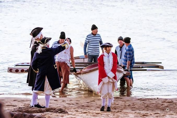 People in period costumes standing by a small boat on a sandy shore near the water