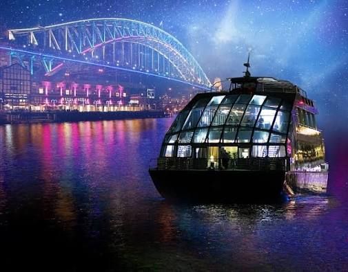 Glass boat on a river at night with a lit bridge and colorful city reflections
