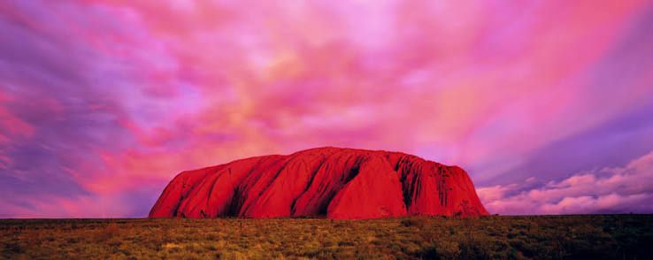 Red rock monolith under a vivid pink and purple sunset sky in a flat desert landscape