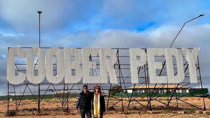 Two people standing before a large “TURKMENBASHY” sign under a blue sky