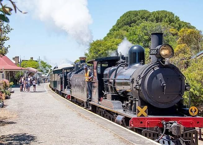 Black steam locomotive at a rural station with passengers and a carriages under a blue sky
