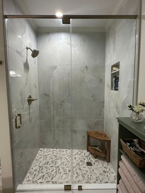 Walk-in shower with glass door, gray tile walls, patterned floor, and small wooden bench