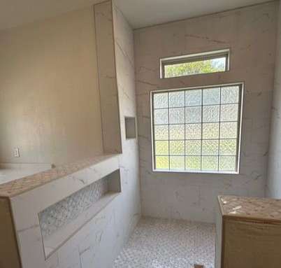 Empty beige-tiled bathroom with a window, built-in shelf, and stone countertop.