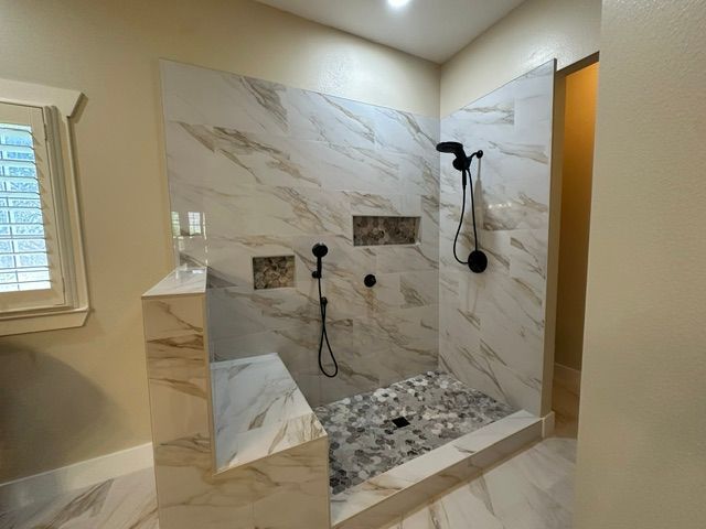Marble-tiled walk-in shower with black fixtures, bench, and pebble floor in a bright bathroom