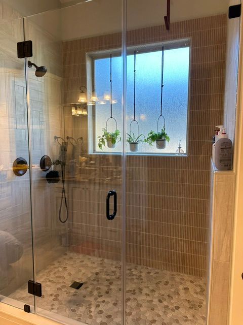 Modern shower with glass enclosure, beige tile walls, mosaic floor, and a frosted window with plants.