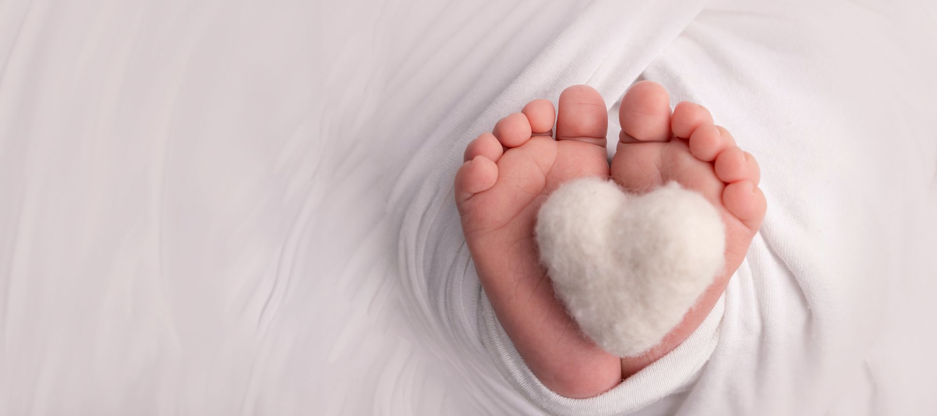 Feet wrapped in white fabric, toes holding a fluffy white heart on a soft pink background