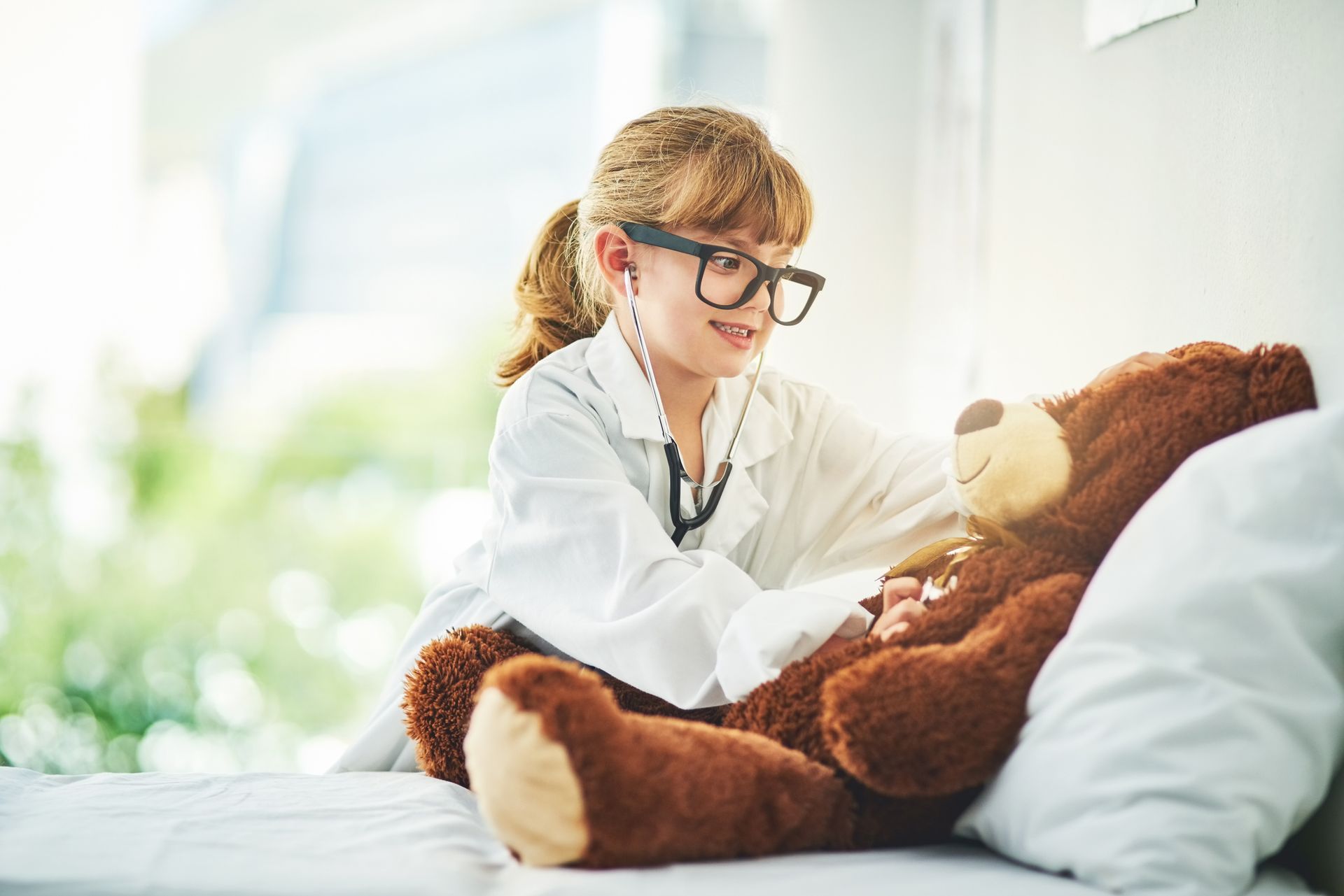 Girl in doctor's coat with stethoscope examining a teddy bear on a bed.