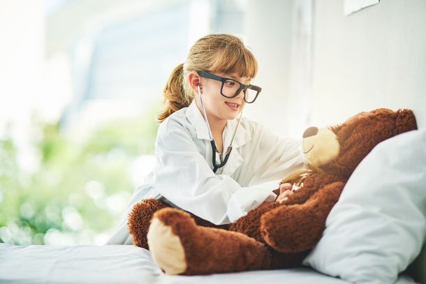 Young child in a white coat and glasses plays with a teddy bear on a bed