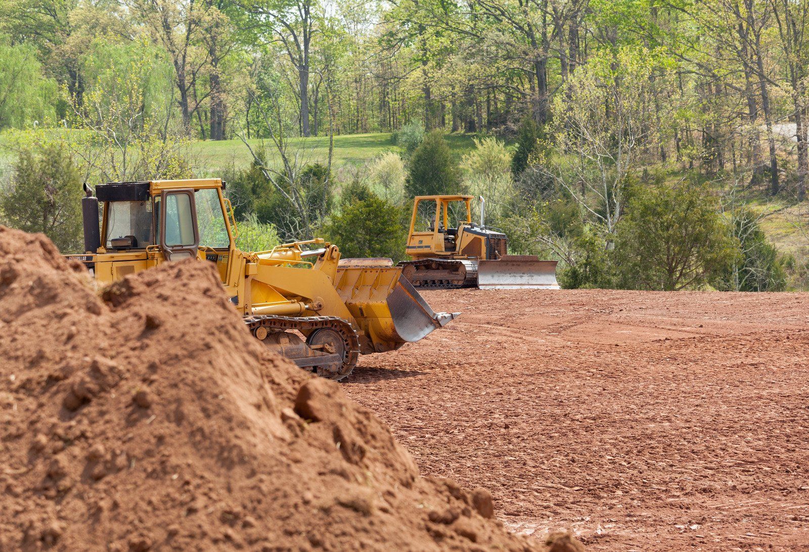 Two yellow bulldozers on a dirt lot, clearing land, with trees in the background.