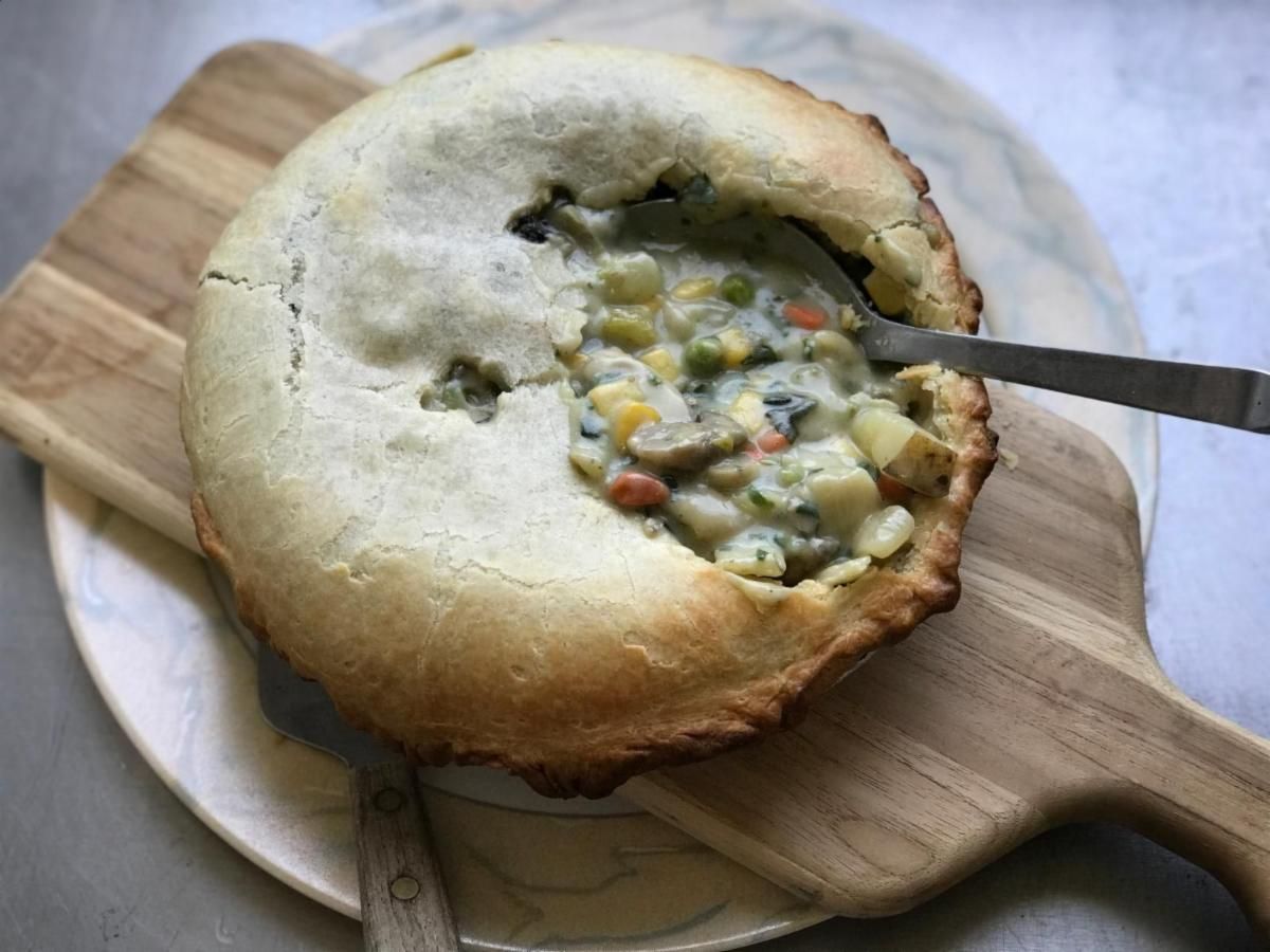 Chicken pot pie, crust partially broken, spoon scooping filling on a wooden board.