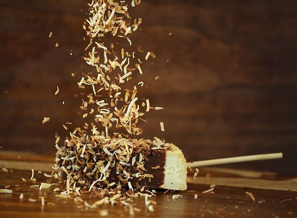 Chocolate-covered banana on a stick with shredded coconut being sprinkled over it, on a wooden surface.
