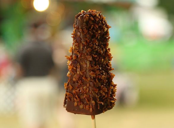 Chocolate-covered frozen banana on a stick, coated with chopped nuts, blurry outdoor background.