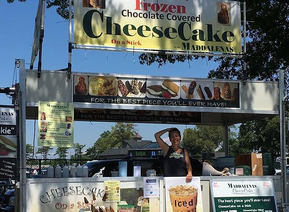 Food vendor selling frozen chocolate-covered cheesecake on a stick. Person behind the counter with iced drink, sunny day.