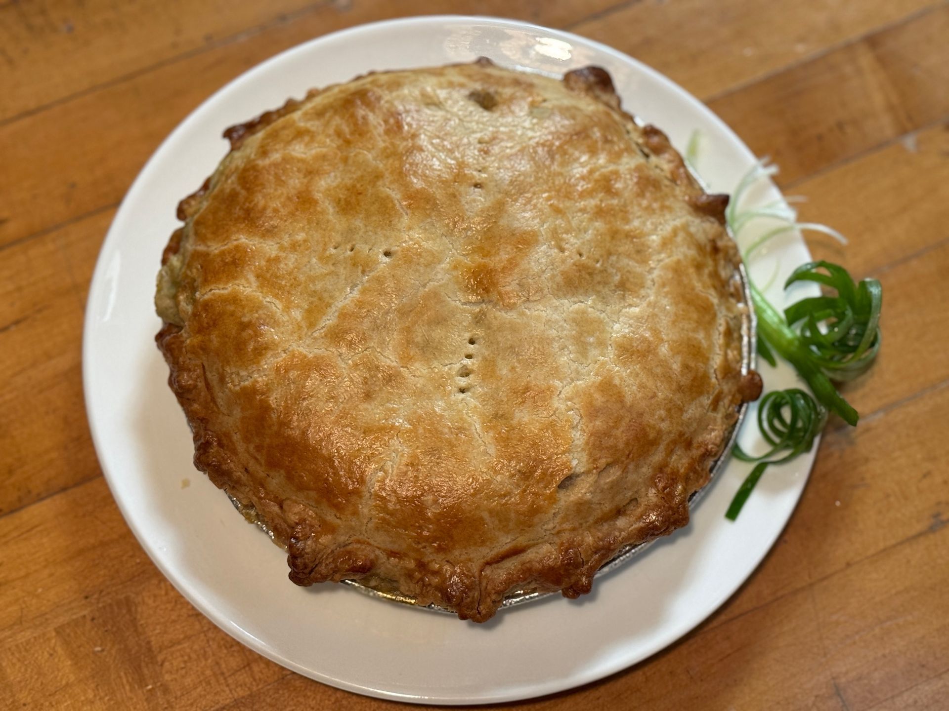 Golden-brown baked pie on a white plate, with green onion garnish and a wooden surface in the background.