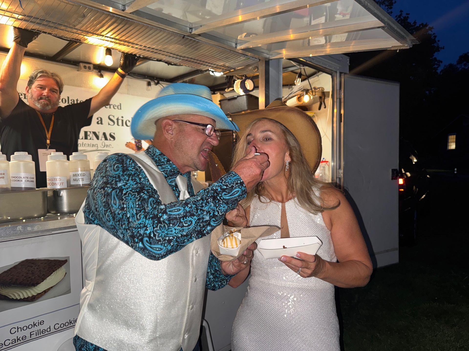 Couple sharing ice cream near a food cart, man in cowboy hat feeding the woman.