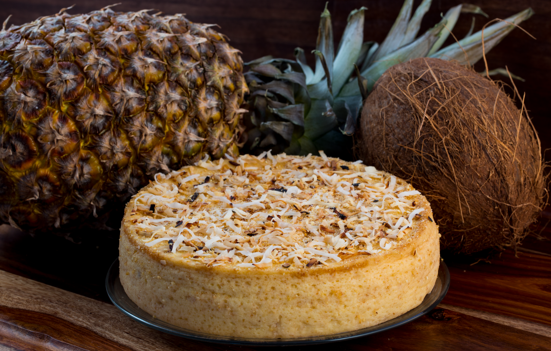 A pineapple and coconut are sitting next to a cake on a wooden table.