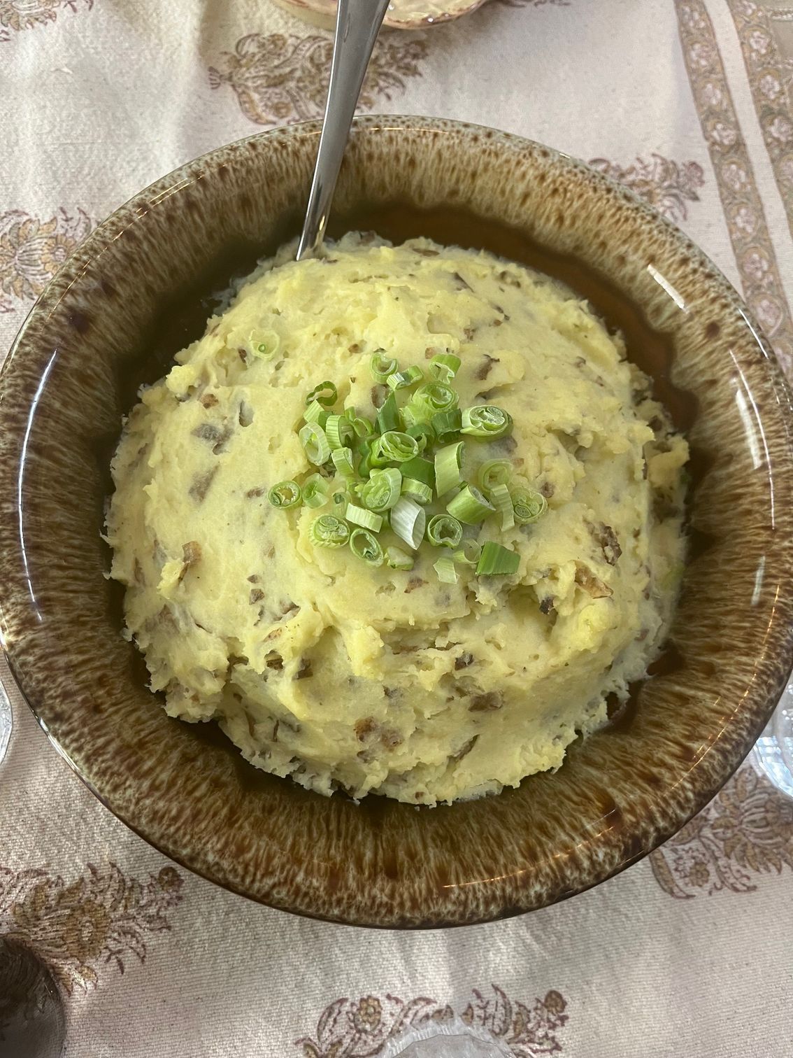 Mashed potatoes with chives in a brown bowl on a patterned surface.