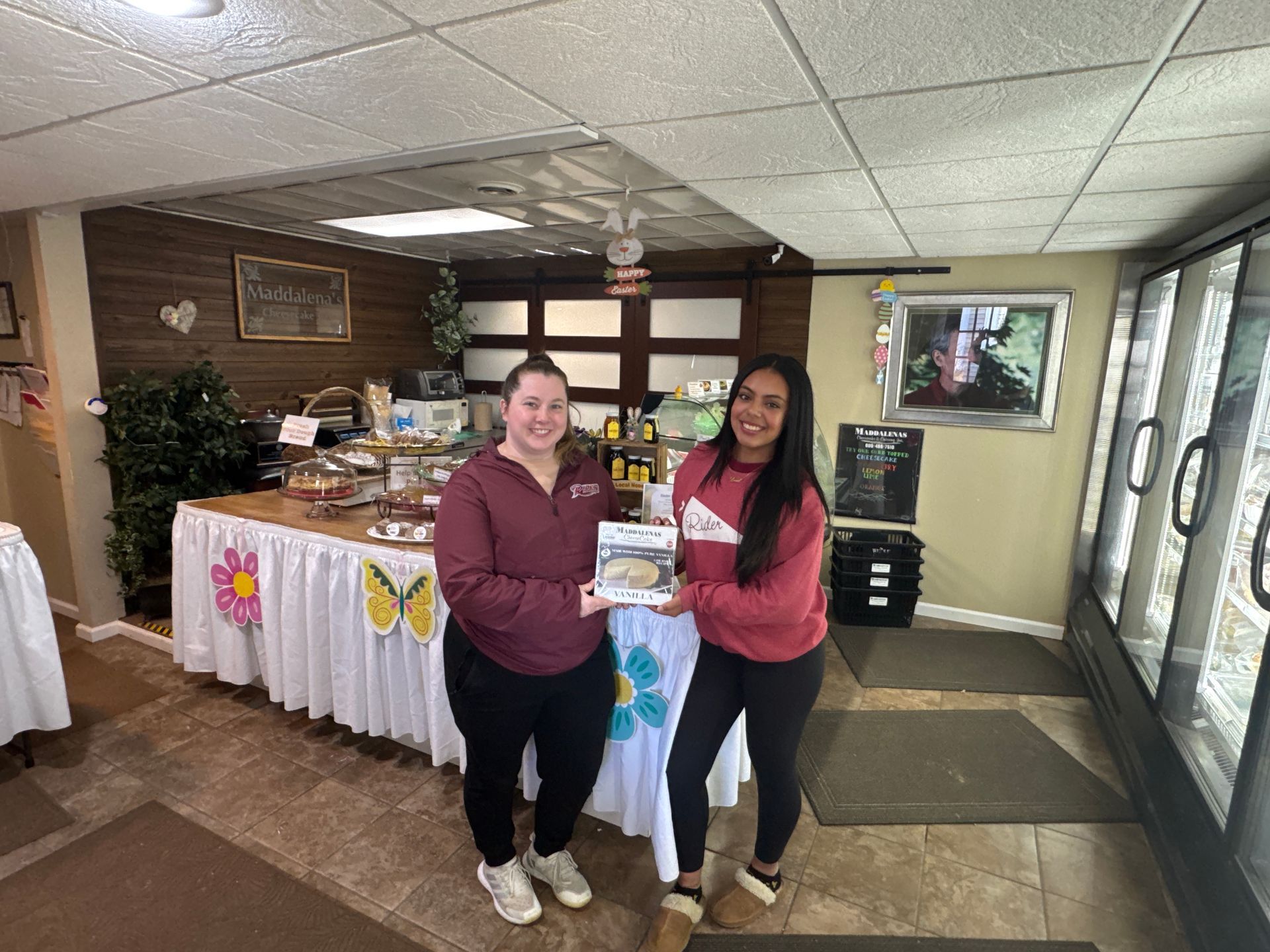 Two women holding a product, standing near a food display table in a shop.
