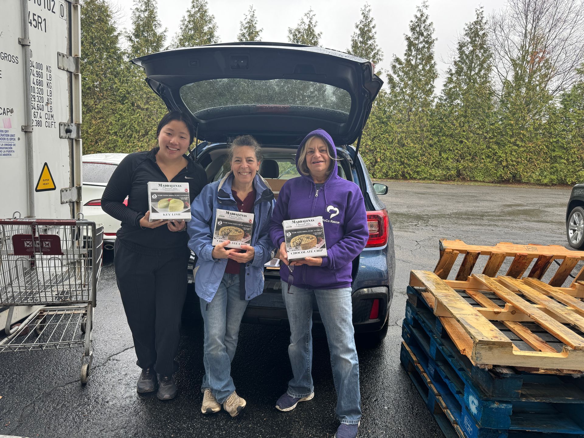 Three people hold food boxes near an open car trunk; pallets and a shopping cart are nearby.
