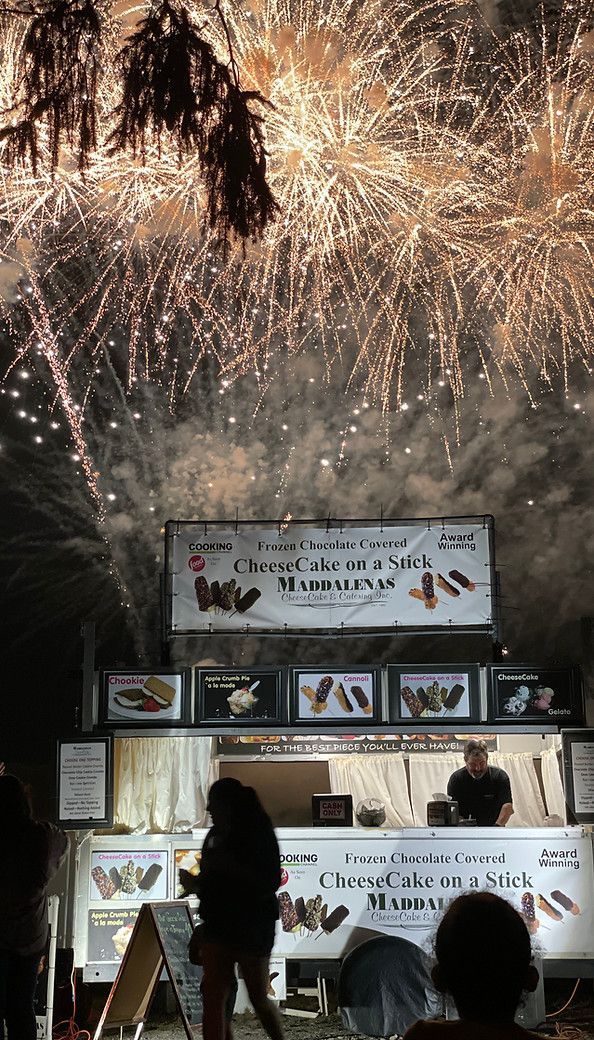 A group of people standing in front of a food stand watching fireworks.