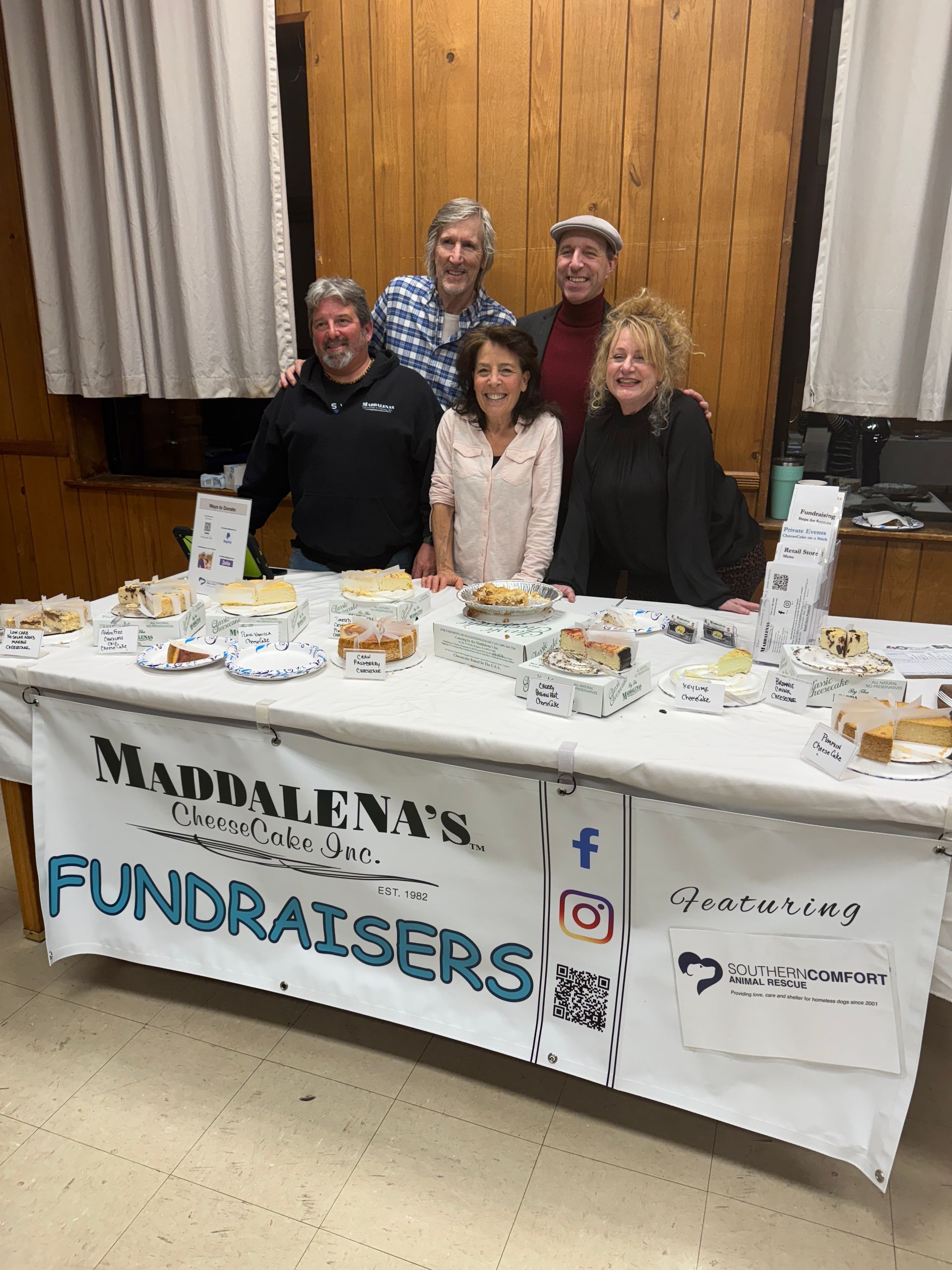 People at a Maddalena's fundraising table display pastries. Table has a banner. Wooden paneling in the background.