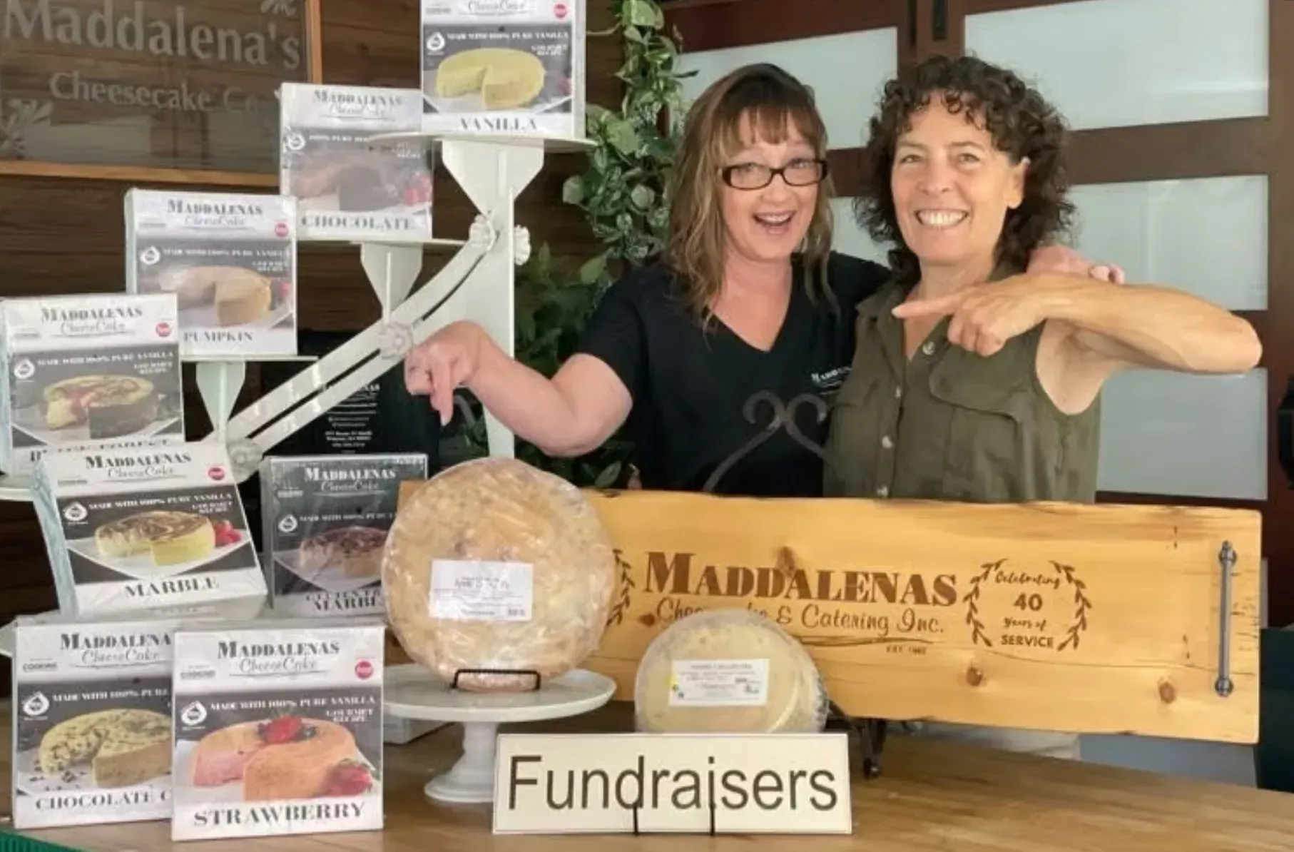 Two women stand behind a display of cheesecakes, pointing and smiling. A sign reads