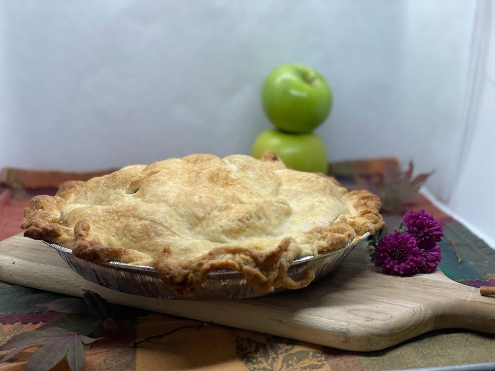 Apple pie on a wooden board with green apples in the background and fall foliage.