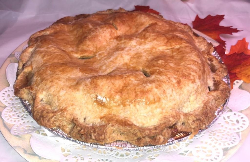 Apple pie in a foil tin, golden crust, on a decorative paper doily with fall leaves.