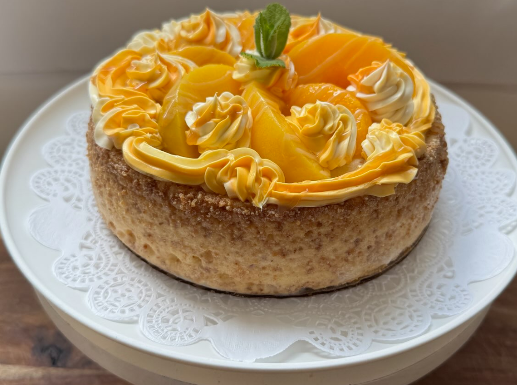 Cheesecake topped with yellow frosting, peach slices, and whipped cream on a cake stand.