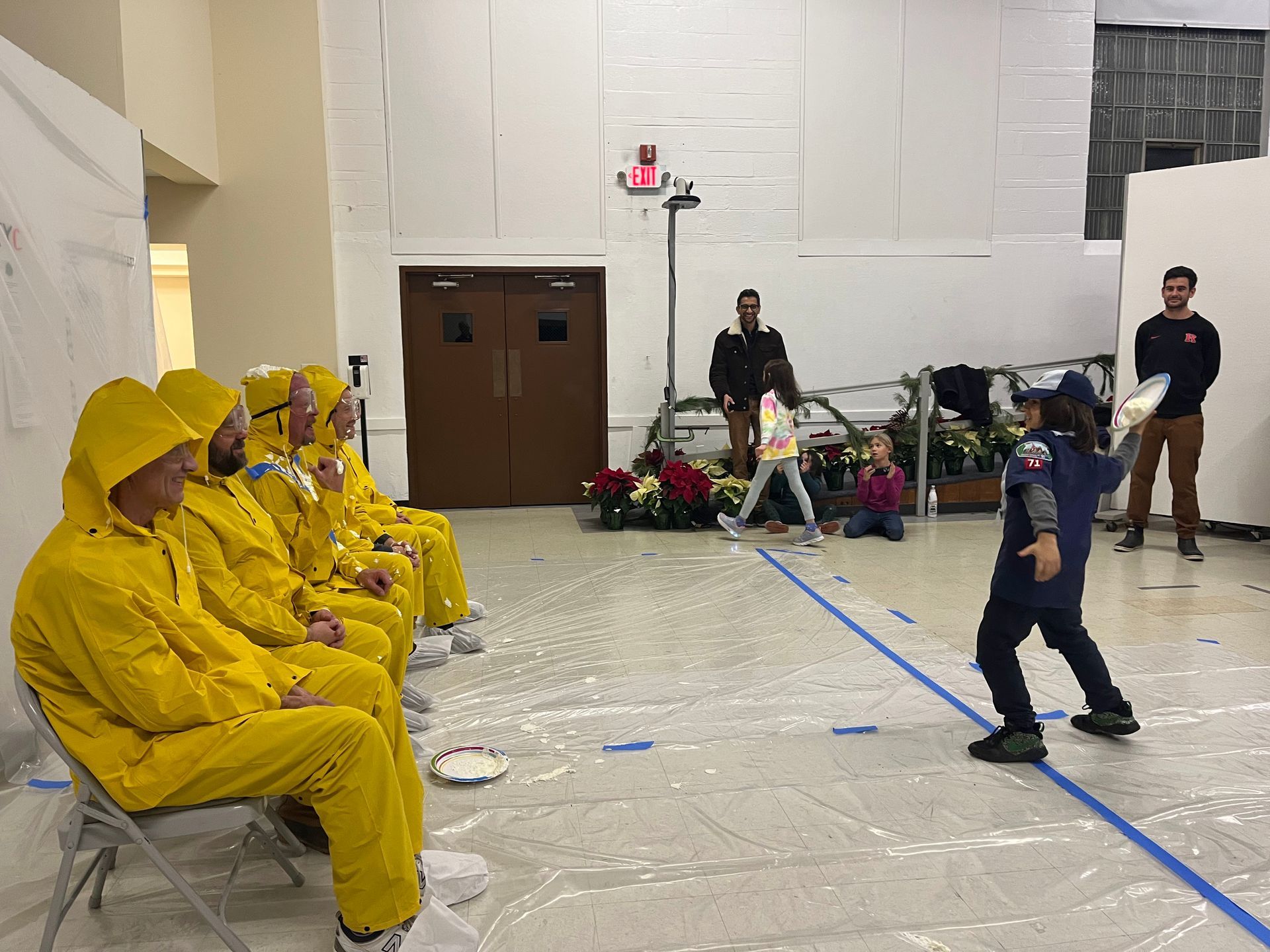 A child throws a pie at people in yellow rubber raincoats suits seated on chairs, inside a hall.