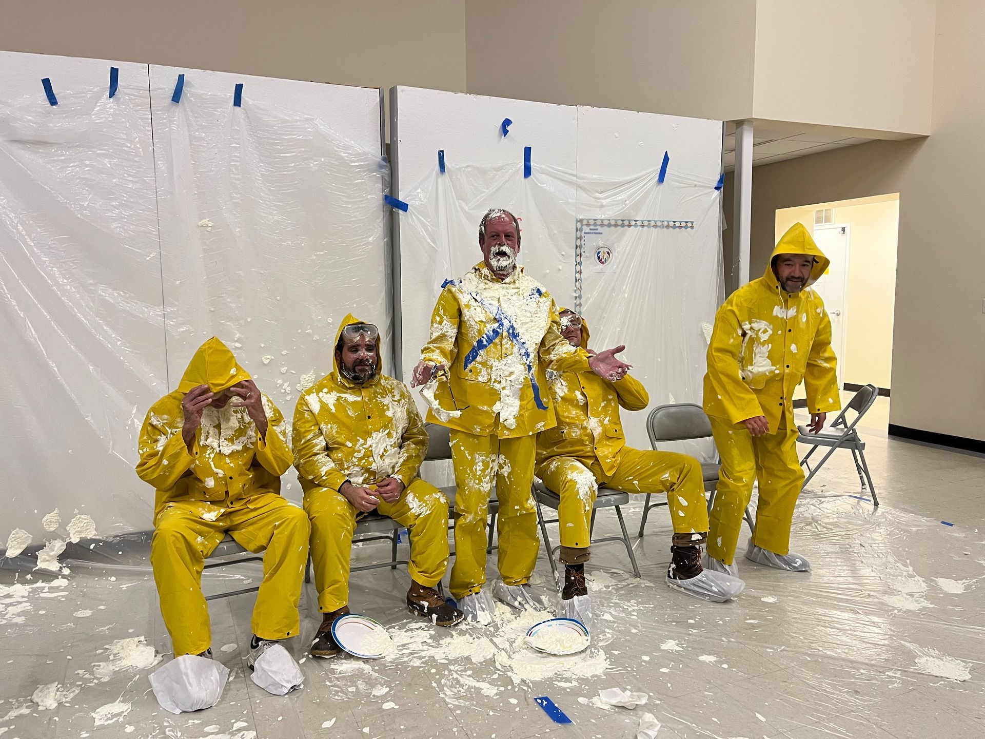 Five people covered in whipped cream and wearing yellow suits pose after a pie-in-the-face event.